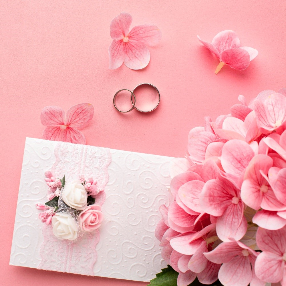 Wedding invitation card with pink flowers and two rings on a soft pink background.